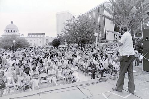 A man stands on an outdoor stage, speaking through a microphone to a large seated crowd, with a domed building in the distance. Black-and-white photo.