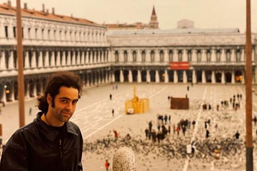 A man with dark curly hair in a ponytail and a  black leather jacket poses, smirking, with the open square of a Gothic-style church behind him. 
