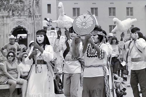 A small group of people wearing white dresses and embroidered shirts process outdoors in front of a crowd. They were white face paint or masks, and two carry papier mache figures of white ducks in flight, and one person holds a white circular emblem.