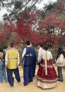 A group of young people stand at the edge of a pond, wearing traditional clothes: the men in shimmering vests over long-sleeve white shirts and loose shimmering pants in navy blue. A woman wears a white blouse, wide marroon skirt with cream-colored embroidery, and a bow tied in the back. On the far side of the pond, leaves on the trees have turned red.