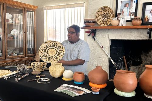 A man holds up a shallow, circular basket with a black flower design. On the table in front of him are more baskets and terra cotta-colored pots.
