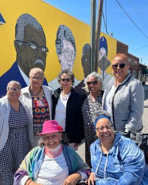 Seven women pose, smiling, in front of the mural with yellow background.