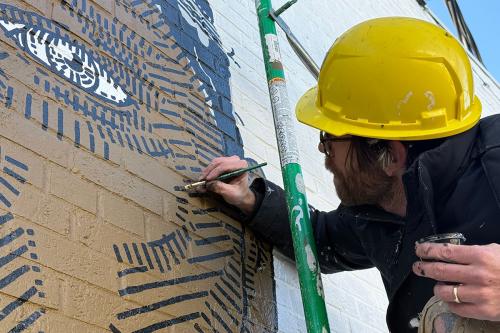 A man wearing a black jacket and yellow hard hat stands on scaffolding white painting detailed black shadow lines on a large portrait.