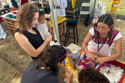 A woman in a floral embroidered top and white apron, with a feather adornment in her hair, sorts through colorful dyed feathers in a plastic bin, set on a table outdoors as festival visitors stand around to watch and make their own feather adornments.