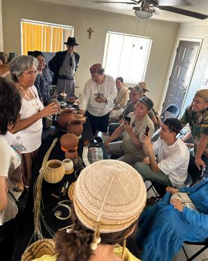 A group of people crowd around a table set with pottery and baskets in a narrow living room.