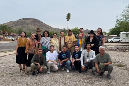 Seventeen people pose, standing and squatting, in a large gravel driveway. In the background is a bare hill, palm trees, and parked cars.