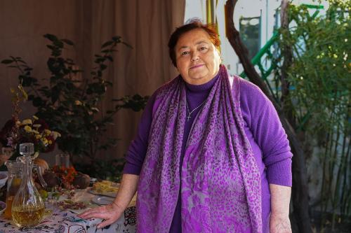 A woman with bright purple sweater and scarf poses next to an outdoor dining table set with drinks and dried flowers.