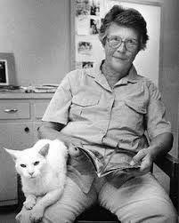 Elder woman sits in a chair, with a magazine and a white cat in her lap. Black-and-white photo.
