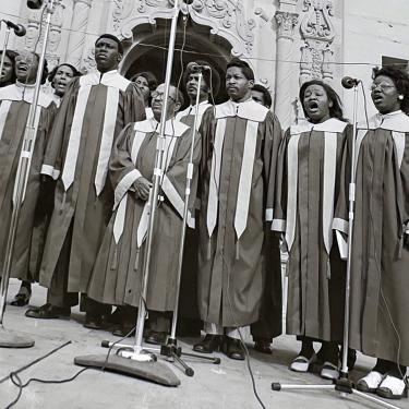 A choir in matching long robes sings on an outdoor stage.