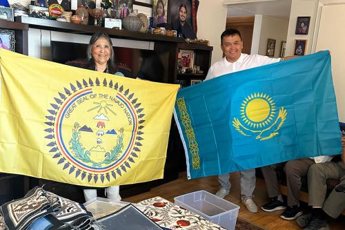 Two people hold up flags: a woman with the yellow Navajo National flag, with symbols of the sun, animals, mountains, and arrowheads, on the left, and a man with the blue Kazakhstan flag, with yellow sun, bird, and decorative side border, on the right.