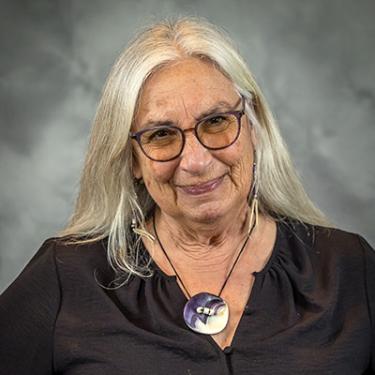 Headshot of Karen Ann Hoffman, with long silver hair, glasses, and a circular shell necklace.