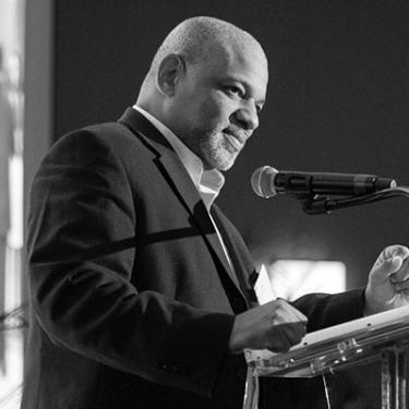 Robert Johnson, wearing a dark suit, stands at a podium with a microphone.