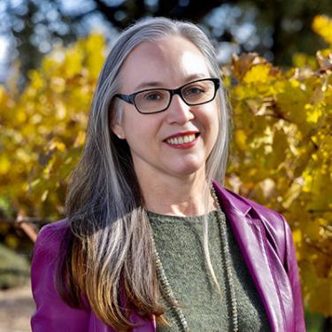 Headshot of Violet Grgich with long silver hair, glasses, and a bright purple jacket, with a vineyard in the background.