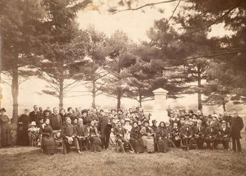 Sepia-toned photo of dozens of people sitting and standing around a white stone memorial column.