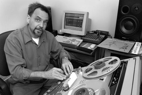 A man smiles as he operates a reel-to-reel tape machine, with a computer and sound equipment behind him.