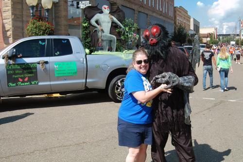 At an outdoor street festival, a woman embraces a person dressed us in a dark fuzzy Mothman costume, with a dark mask with red eyes and lips.