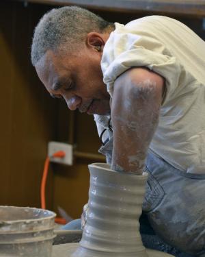 An older Black man at a pottery wheel, one fist down the center of a cylinder of clay.