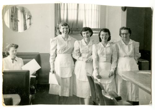 Four women in a diner, wearing matching waitress uniforms and aprons, all smiling. Black-and-white photo.