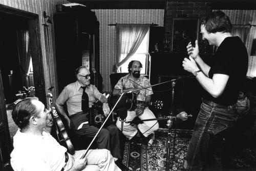 Three men seated in in front of microphones in a living room, holding a fiddle, accordion, and acoustic guitar. They look up at Chris, standing at the right, who is gesticulating at them.