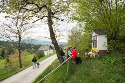 A few people sit at a bench in front of a small outdoor shrine on a hillside above a narrow road. Bouquets of flowers set in front of the shrine.