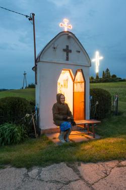 At dusk, someone sits outside a roadside shrine warmly lit from within. Two crosses on the outside glow against the dark sky.