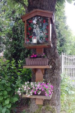 A shrine built onto the trunk of a tree, with three tiers: a display of Mary in the top, and two shelves of flower pots below.