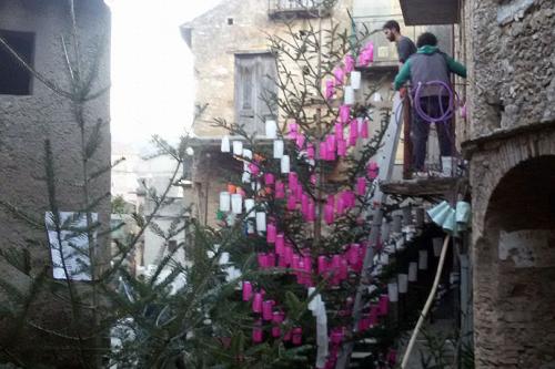 Outdoors, large fir trees are stood up next to stone buildings. Men a second-floor balcony hand bright pink lanterns on the trees.
