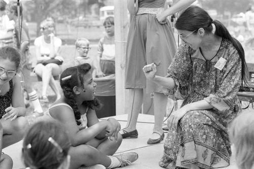 A woman sits, surrounded by young children. She is holding her right hand up in a fist.