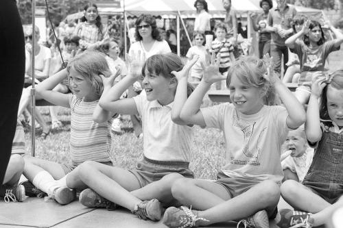 A row of young kids sit on the ground, cross-legged, holding up their hands with splayed fingers to their heads, mimicking an ASL phrase.