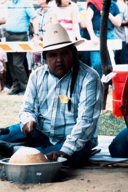 A man, seated on the ground, plays a gourd drum placed in a metal bucket.
