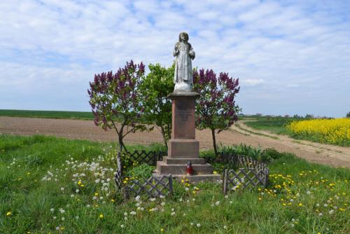 A statue shrine next to a dirt road, surrounded by green grass and wildflowers.