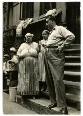 A tall man, in slacks and long-sleeve shirt, stands on the last step of an apartment stoop, holding a cigarette and looking off to the side. A woman next to him looks straight in the camera. Black-and-white photo.
