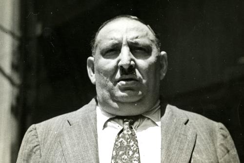 A man  in coat and tie faces the camera, looking just above it, squinting in the sunlight.