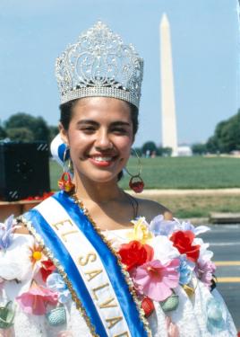 A woman poses smiling with the Washington Monument in the background. She wears a crown, a sash with the words El Salvador, and a white blouse with colorful floral detail