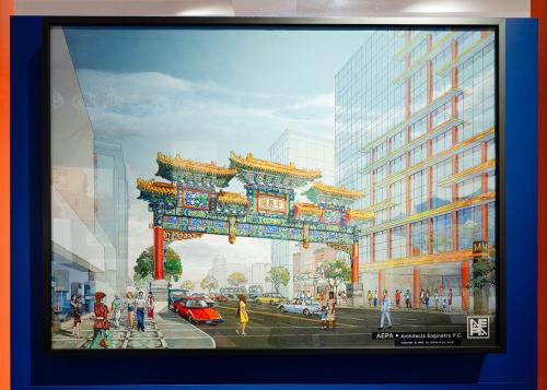Museum display of a full-color architectural drawing of a Chinese-style archway over a city street. The arch has red pillars and decorative blue, green, red, and yellow carving across the top.