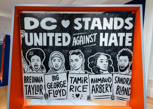 Museum display of a black-and-white poster with bold block letters across the top: DC Stands United Against Hate. Below, five people hold up signs each reading a name: Breonna Taylor, Big George Floyd, Tamir Rice, Ahmaud Arbery, Sandra  Bland.