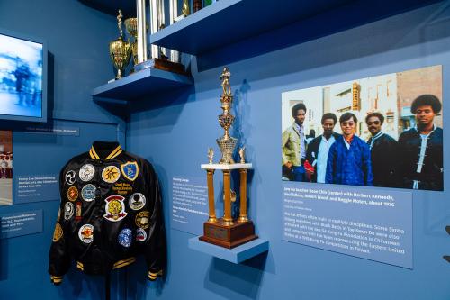 A corner of a museum gallery room displaying martial arts memorabilia: a jacket adorned with several patches, a trophy, an enlarged photograph, and a video screen.