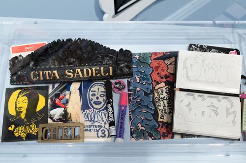 Museum case displaying a sketchbook with graffiti lettering in pencil, illustrations in pen, two markers, a sticker, a photographer of the artist painting, a wooden carving with a floral design and the name Cita Sadelia painted in gold, and a bronze belt 