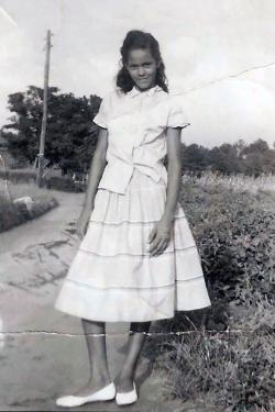 A teenage girl poses for a photo on a rural road. The black-and-white photo has been crinkled and stained.