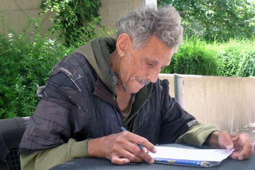 Alexander, with longer hair and warmer clothes, sits at an outdoor table reviewing a document with pen in hand.