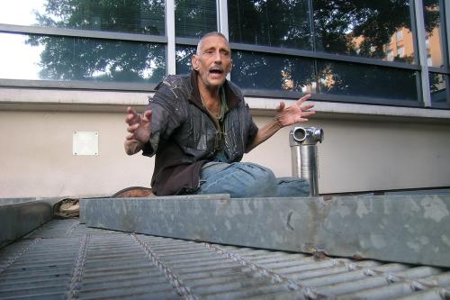 A wiry man in tattered clothes gesticulates passionately as he speaks, sitting atop an outdoor heating grate.