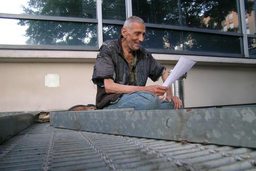 A wiry man in tattered clothes smiles as he reads an article on white printed paper.