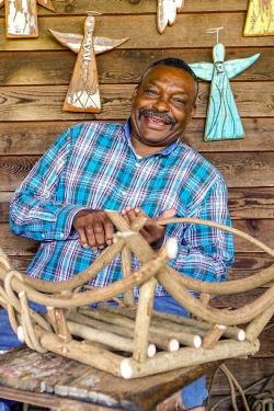 An older man wearing a blue, purple, and white plaid button-up shirt and jeans works a branch in front of a basket currently in development. He appears in front of a wooden background with orange, yellow, and blue angel figures with halos.