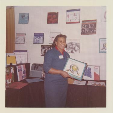 A woman with dark skin and long blue dress holds up a twelve-inch record. More records are on display behind her on a table and the wall.