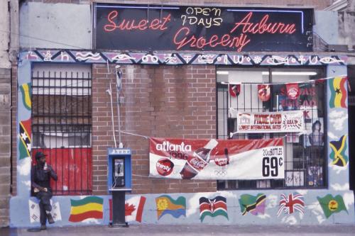 Grocery storefront, with a sign above the window: Sweet Auburn Grocery. Around the window is a mural of various national flags. In the window is an promotion for Coca-Cola during the 1996 Olympics. A man sits in the window.