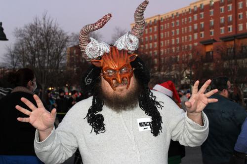 A person a normal long-sleeve T-shirt but also a red Krampus mask with jagged horns. He holds out his hands, fingers splayed, as if to scare onlookers.