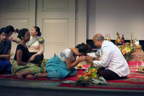 Performers sit casually, cross-legged on stage. A woman bows her head to a man, holding his hands to her forehead.