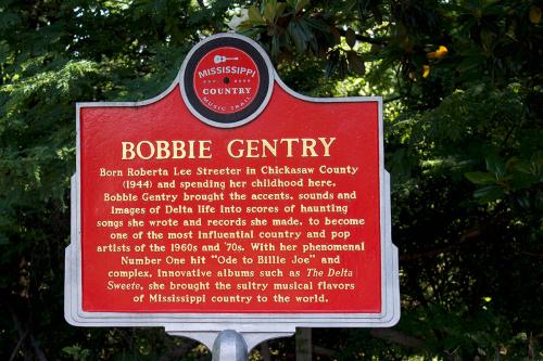Red sign marker on the Mississippi Country Music Trail. The signs reads: Born Roberta Lee Streeter in Chicksaw County (1944) and spending her childhood here, Bobbie Gentry brought the accents, sounds and images of Delta life into scores of haunting songs 