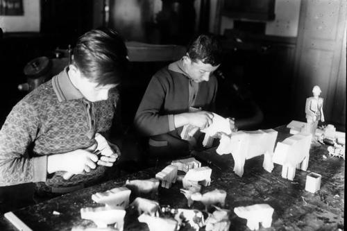 Two young people carve wooden cow figurines. Black-and-white photo.