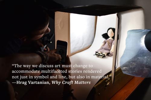 A man is bent over to look into a handmade light box containing a small, handmade cloth doll. In front of the light box, a person holds a phone to capture a photo of the doll. A quote is overlaid: “The way we discuss art must change to accommodate multifa